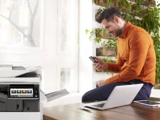 Man using laptop and mobile phone with a printer in foreground